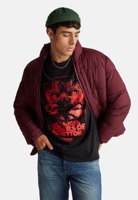 Chaqueta acolchada color granate con textura acolchada, usada sobre una camiseta negra con diseño gráfico rojo. Combinada con jeans azules y un gorro verde.