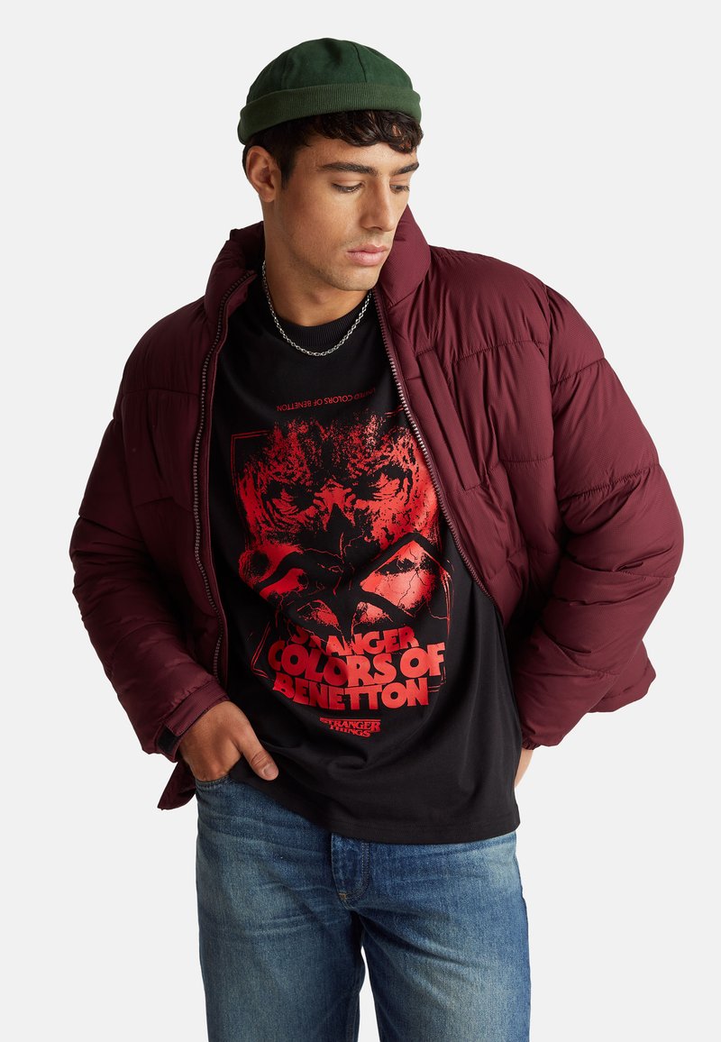 Chaqueta acolchada color granate con textura acolchada, usada sobre una camiseta negra con diseño gráfico rojo. Combinada con jeans azules y un gorro verde.