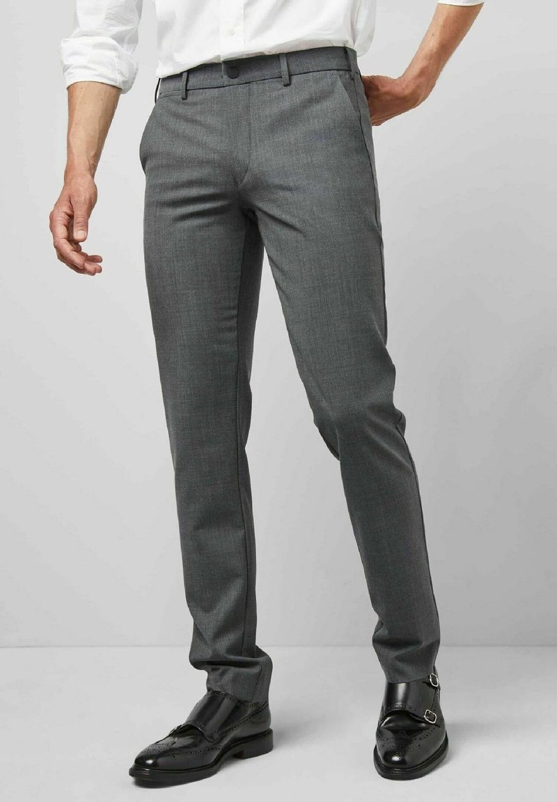 Homme portant un pantalon de costume gris slim fit, une chemise blanche et des chaussures en cuir noires à double boucle monk, debout sur un sol gris.