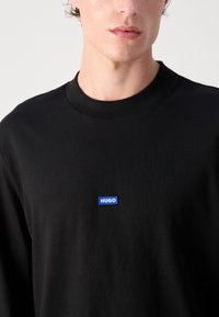Zwart langslavende shirt met ronde hals, voorzien van een klein blauw "HUGO" logo patch op de borst. Glad katoenstof.