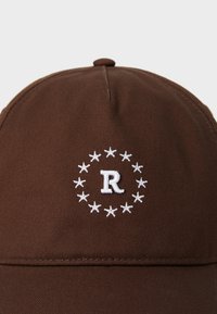 Pruun kangast müts, mille ees on valgest tikitud täht "R", mida ümbritseb ringikujuline valgete tähtede muster.