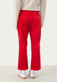 Pantaloni a zampa d'elefante in corduroy rosso con due tasche posteriori, abbinati a un maglione beige chiaro e scarpe da ginnastica bianche, mostrando un tessuto testurizzato.