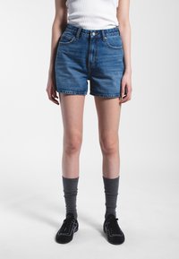 Denim shorts in medium blue featuring a high waist, five-pocket design, and frayed hem. Worn with gray knee-high socks and black shoes.