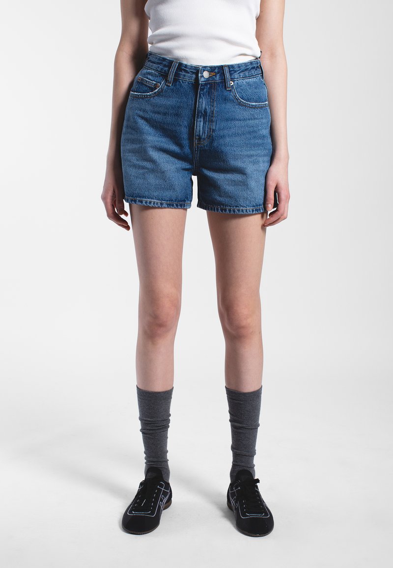 Denim shorts in medium blue featuring a high waist, five-pocket design, and frayed hem. Worn with gray knee-high socks and black shoes.