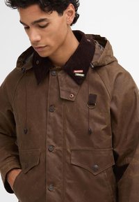 Brown waxed jacket with a corduroy collar, snap buttons, and drawstring hood. Features two front pockets and brand labels on the collar.