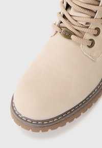 Botín de tobillo de ante beige con punta redonda, cordones perforados y ojales de metal, adornado con costuras decorativas a lo largo de la suela de goma.