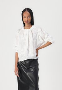 Hvit brodert bluse med samlet halslinning og lange, puffete ermer, sammen med et elegant svart skinnskjørt. Teksturert blomsterdesign er synlig.