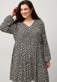 Schwarzes Kleid mit einem weißen Blattmuster, V-Ausschnitt, langen Ärmeln und gerafftem Bund, aus leichtem Stoff gefertigt.