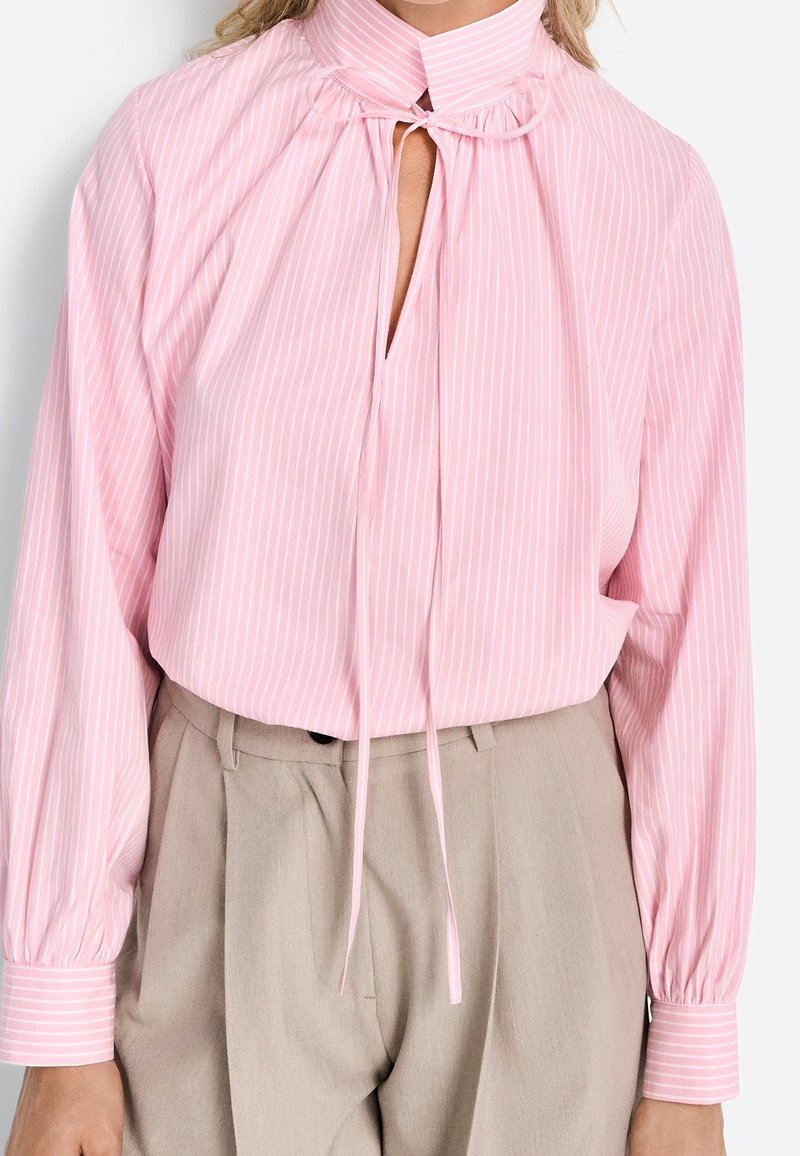 Hemdbluse in zartem Rosa mit langen Ärmeln und vertikalen weißen Streifen, hohem Kragen mit Bindebändern, gathered Details und geknöpften Ärmeln, kombiniert mit beigen Hosen.
