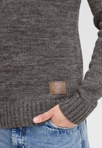 Grauer Strickpullover mit einem gerippten Saum und Bündchen, versehen mit einem Lederetikett mit der Aufschrift "Solid." Die Textur ist grob und gemütlich.