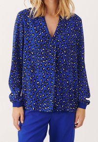 Femme portant un chemisier à manches longues bleu cobalt avec un imprimé léopard noir et blanc et un pantalon bleu assorti, debout devant un fond uni.