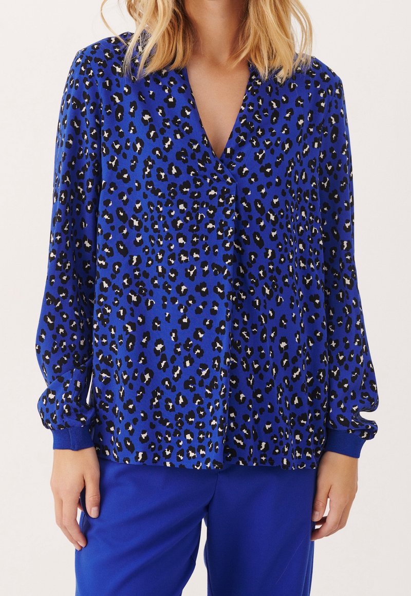 Femme portant un chemisier à manches longues bleu cobalt avec un imprimé léopard noir et blanc et un pantalon bleu assorti, debout devant un fond uni.