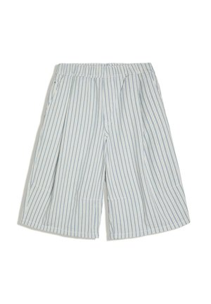 Homme appuyé contre un comptoir carrelé jaune, portant une chemise blanche à manches courtes, un short large rayé, des chaussettes blanches et des mocassins noirs.