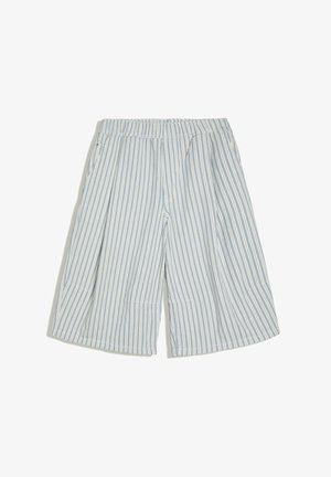 Man leunt tegen een geel betegelde toonbank met een wit kortemouwshirt, gestreepte wijde shorts, witte sokken en zwarte loafers aan.