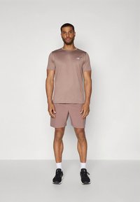 Conjunto atlético masculino composto por uma t-shirt de manga curta ajustada, de cor castanho claro, e calções combinando, feitos de tecido macio. Ténis de corrida pretos com detalhes brancos.