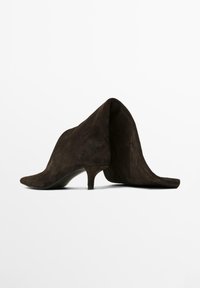 Un paio di stivaletti in suede marrone scuro con punta affusolata e tacco corto a kitten, mostrati su uno sfondo bianco.