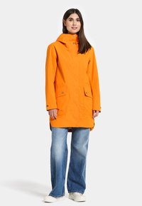 Orange vattentålig parkas med huva, två framfickor, tryckknappar och knälång design, kombinerad med vida blå jeans.