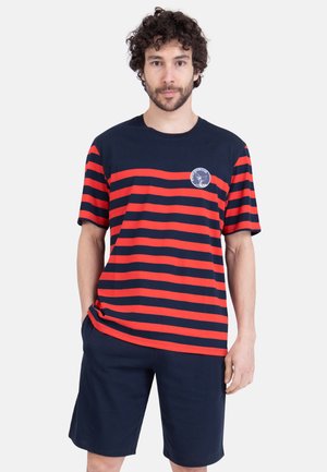 Homme portant un t-shirt à manches courtes rayé bleu marine et rouge avec un logo circulaire, associé à un short bleu marine, debout les mains dans les poches.