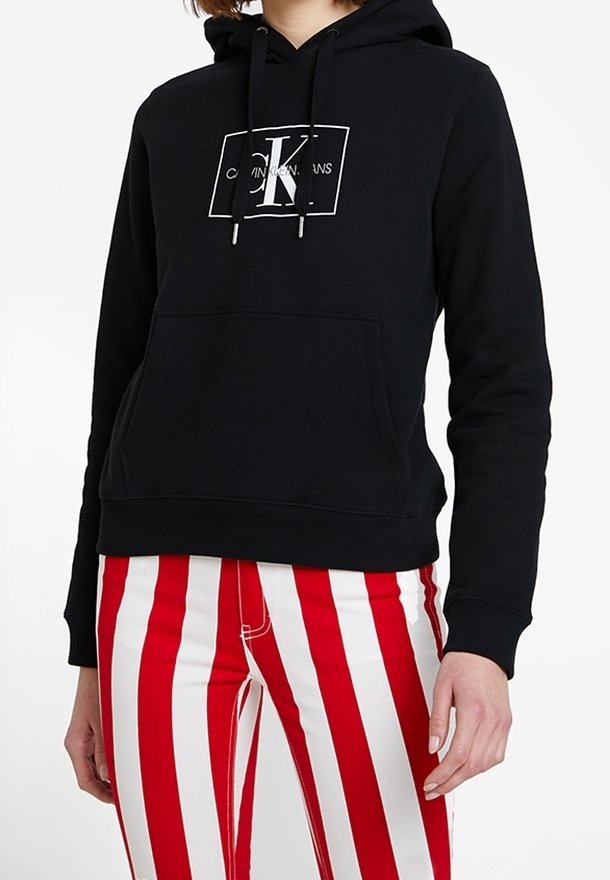 Sudadera negra con capucha y bolsillo canguro, con el logo blanco de "Calvin Klein". Combinada con pantalones a rayas verticales rojas y blancas.