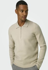 Beiger Strickpullover mit einem Reißverschlusskragen, langen Ärmeln und einer glatten Textur, präsentiert an einem Model vor einem hellen Hintergrund.