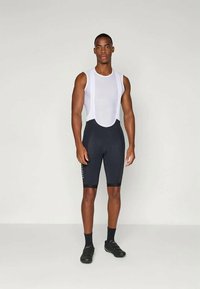 Sportos férfi áll fekete kerékpáros rövidnadrágban, fehér hálós ujjatlan felsőben, fekete zokniban és fekete kerékpáros cipőben egy egyszínű háttér előtt.