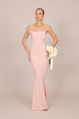 Žena u blijedo ružičastoj, uskoj haljini bez rukava s cvjetnim detaljem, drži bijele kale, nosi zlatni nakit, pozira na bež pozadini.