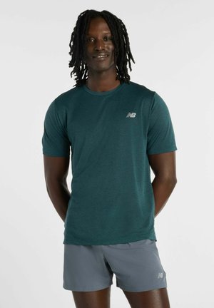 Mann mit Dreadlocks, trägt ein kurzärmeliges türkises New Balance-Shirt und graue Sportshorts, steht mit den Händen hinter dem Rücken.