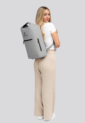 Zaino impermeabile grigio con chiusura a rotolo, tasca con zip laterale e dettagli neri, indossato sulla schiena da un modello in pantaloni chiari.
