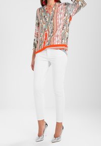 Colorful patterned blouse with long sleeves, V-neckline, and orange accents, paired with fitted white pants and silver heels.