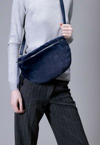 Navyfarbene Tasche aus Kunstfell für die Schulter mit einem metallischen Reißverschluss, halbkreisförmiger Form und einem abnehmbaren Gurt. Die glatte Textur kontrastiert mit der grauen Kleidung.