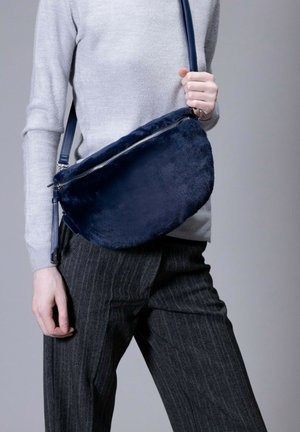Navy faux-fur crossbody bag with a metallic zipper, semi-circular shape, and a detachable strap. Smooth texture contrasts with the grey attire.