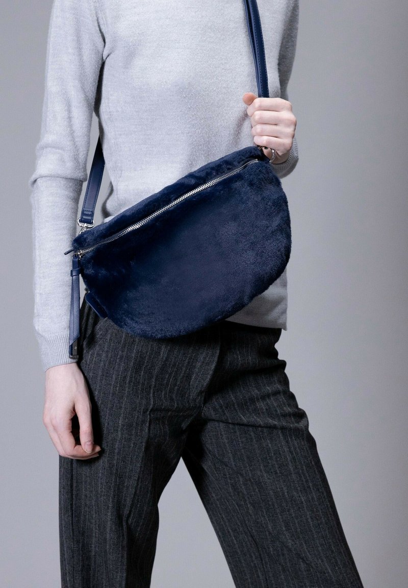 Navyfarbene Tasche aus Kunstfell für die Schulter mit einem metallischen Reißverschluss, halbkreisförmiger Form und einem abnehmbaren Gurt. Die glatte Textur kontrastiert mit der grauen Kleidung.