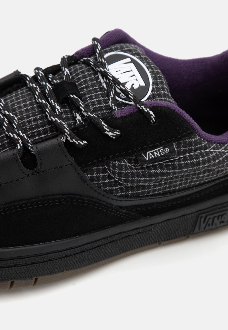 Schwarze Vans Sneaker mit einem karierten Obermaterial in Kombination mit lila Akzenten, schwarzen Wildlederdetails und kontrastierenden weißen Schnürsenkeln und Logo.