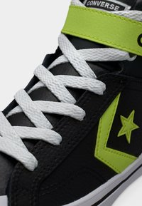 Μαύρο δερμάτινο αθλητικό παπούτσι Converse με λευκά κορδόνια, φωτεινό κίτρινο αστέρι και λογότυπο κεραυνού, φωτεινό κίτρινο λουρί και λευκή καουτσούκ σόλα.