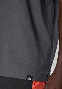 Lähi kuva henkilöstä, joka käyttää tummaa harmaata Adidas-paitaa, jossa on logotagi helman lähellä, sekä mustia ja punaisia urheilushortseja.