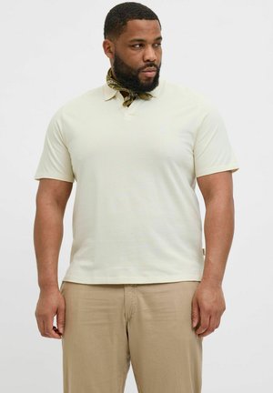 Lichtbeige poloshirt met korte mouwen, ribbelstructuur en een twee-knops sluiting. Een paar beige broeken en een gestructureerde sjaal om de nek.