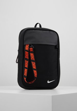 Kompaktowa czarna torba Nike z przednią kieszenią, czerwoną paskiem z logo Nike oraz białym logo Nike swoosh na jednolitym szarym tle.