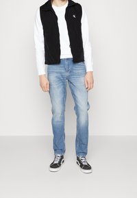 Czarna pikowana kamizelka z wysokim kołnierzem na białej koszuli z długim rękawem, połączona z jasnoniebieskimi dżinsami i czarnymi trampkami.