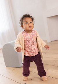 Helles beigefarbenes Cardigan über einem pinkgemusterten Oberteil und dunkelvioletten Cordhosen. Die Schuhe sind hellbraun mit durchbrochenen Akzenten.