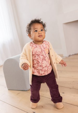 Helles beigefarbenes Cardigan über einem pinkgemusterten Oberteil und dunkelvioletten Cordhosen. Die Schuhe sind hellbraun mit durchbrochenen Akzenten.