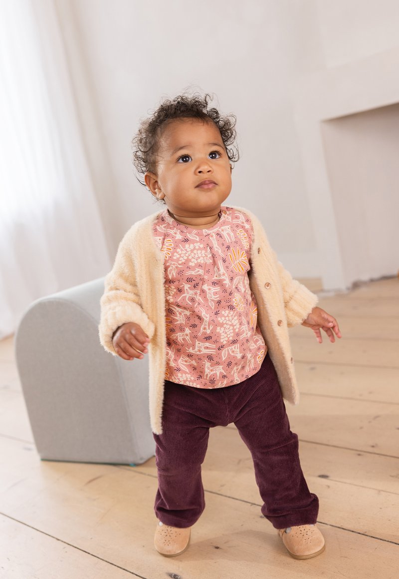 Helles beigefarbenes Cardigan über einem pinkgemusterten Oberteil und dunkelvioletten Cordhosen. Die Schuhe sind hellbraun mit durchbrochenen Akzenten.
