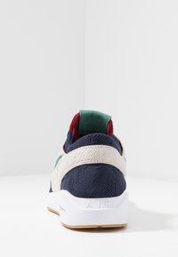 Baskets en maille marine avec des accents crème et verts, présentant une surface texturée et une semelle blanche. Le talon affiche "AIR" en lettres en relief.