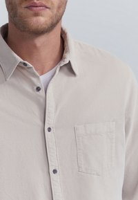 Camisa de corduroy beige claro con cierre de botones en la parte delantera, cuello puntiagudo y un bolsillo en el pecho, que presenta botones oscuros y una textura de ribeteado vertical.