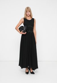 Crna maksi haljina s prilagođenim bodicem, ukrašena malim zakovicama, lepršava suknja i prozirni rub. Uparena s crnom clutch torbicom i potpeticama.