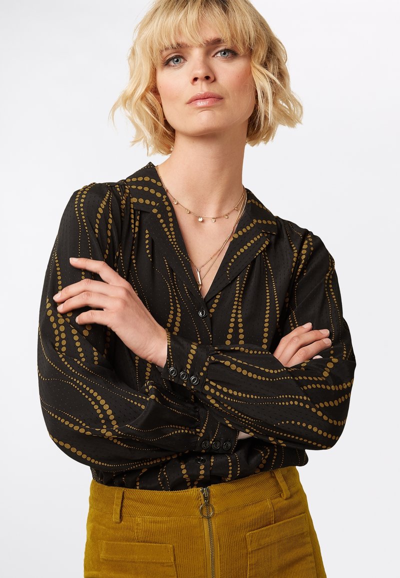 Blusa nera con motivo a pois dorati, polsini abbottonati e colletto. Indossata con pantaloni di velluto a coste color senape con tasche frontali e chiusura con zip.