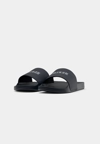 Ein Paar schwarze Rutschsandalen mit breiten Riemen, die auf der Oberseite weißen "Hilfiger"-Text zeigen, vor einem schlichten weißen Hintergrund präsentiert.