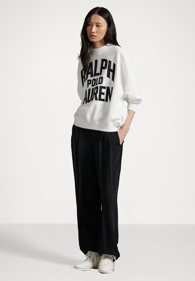 Biela bavlnená mikina s výrazným čiernym textom "RALPH LAUREN", skombinovaná s čiernymi plisovanými nohavicami a bielymi teniskami. Mäkká textúra, voľný strih.