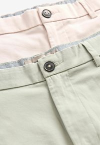 Deux paires de pantalons exposées, l'une rose clair et l'autre verte clair. Les deux comportent des boutons, des passants de ceinture et une texture en coton lisse.