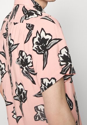 Mężczyzna ubrany w różową koszulę z krótkim rękawem w duże czarno-białe kwiatowe wzory, widziany od tyłu i z boku.