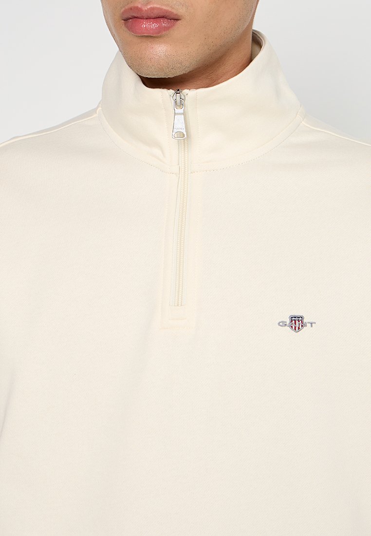Mann in einem cremefarbenen Viertelreißverschluss-Sweatshirt mit kleinem GANT-Logo auf der linken Brust, sichtbar von den Schultern bis zu den Lippen.